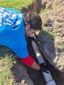 A plumber working on a sewer line repair in a trench, wearing a blue shirt and gloves.